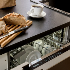 Brot auf Holzbrett mit Brotmesser auf Arbeitsplatte in der Küche, unterhalb der geöffnete Geschirrspüler