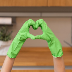 Hände in grünen Gummihandschuhen formen ein Herz vor dem Hintergrund einer Küche mit Holzoberflächen