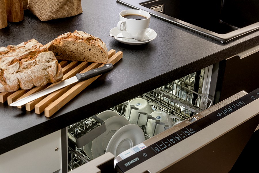 Küchenarbeitsplatte mit Holzbrett, Brot und Brotmesser. Darunter der halb geöffnete Geschirrspüler.